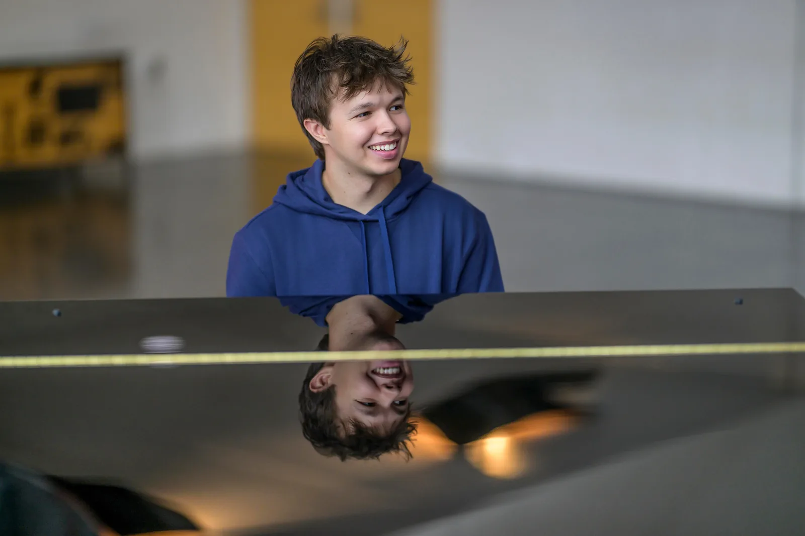 Max Cooper III in a blue hoodie smiling above the piano lid that reflects him